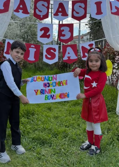 İki öğrencimiz 23 Nisan süslemelerinin önünde 'Bugünün çocukları yarının büyükleri' yazılı pankartı tutuyor.