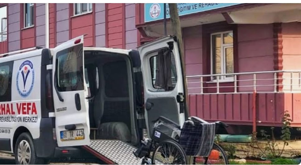 Servis aracının arka rampası açık; yanında tekerlekli sandalye duruyor, arkada merkezin girişi görünüyor.